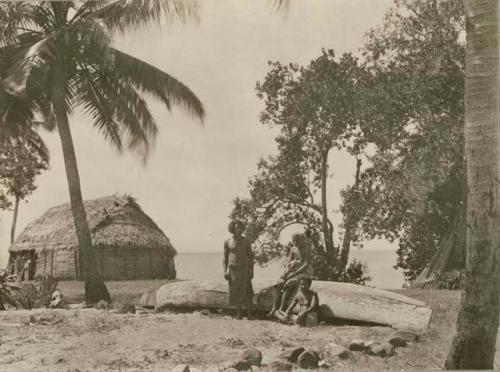 Three men by a canoe, a house and the ocean are visible in the background