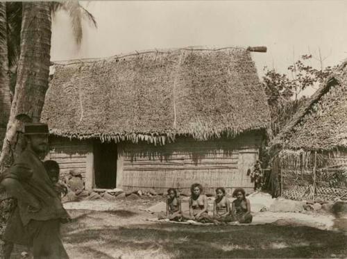 Four native women sitting in front of house, a member of the expedition stands in the foreground to the left