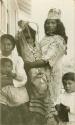 Thompson River Indian women "TsEstaz. tko," in costume with baby carrier, and "Taxinek" with children