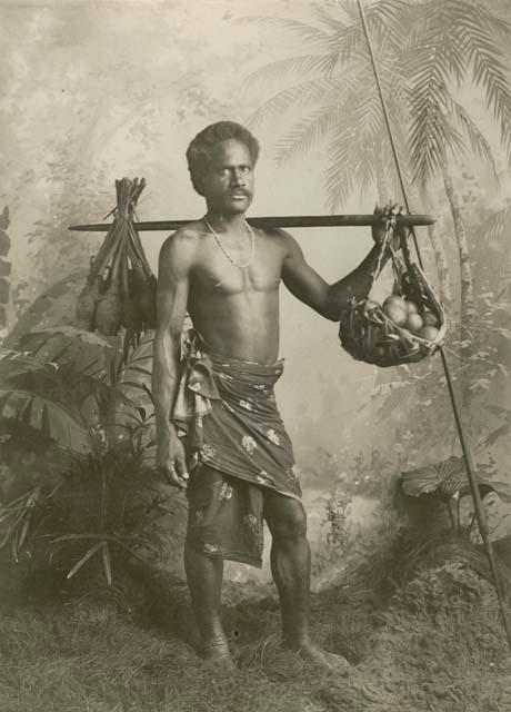 Man in a studio-staged jungle scene carrying fruit on a carrying pole