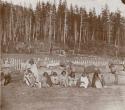 Eight men, women, children sitting for picture; legs, picket fence, forest in ba