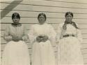 Three women; Upper Lake weavers, standing against wood slat wall
