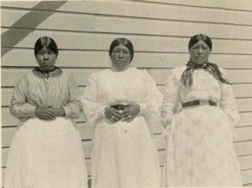 Three women; Upper Lake weavers, standing against wood slat wall