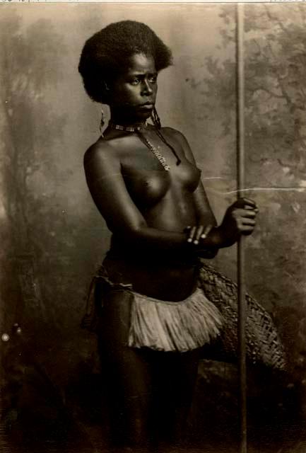 Studio portrait of a Fijian woman holding a spear, carrying a basket, and wearing a grass skirt