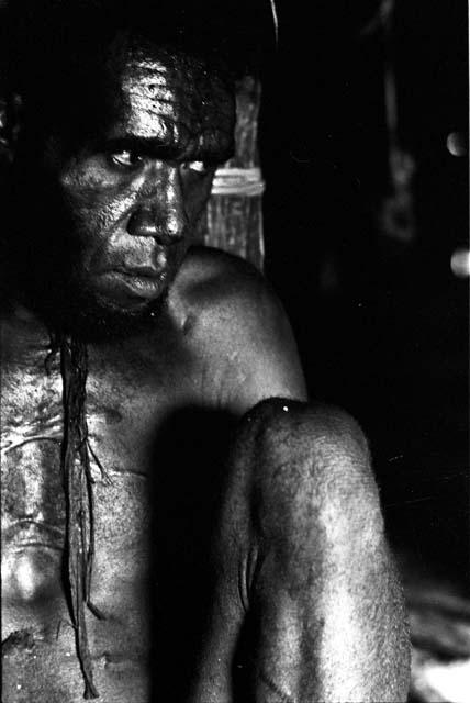 Tuessiké sitting in honai against fireplace, rat hanging behind him