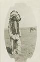 A Jicarilla Apache man wearing a large feather headdress, a horse stands in the background