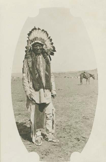 A Jicarilla Apache man wearing a large feather headdress, a horse stands in the background