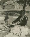 Navajo woman and child shucking corn, with hogan in background
