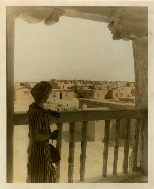 Tinted photograph of a woman in long striped skirt looking over balcony at pueblos