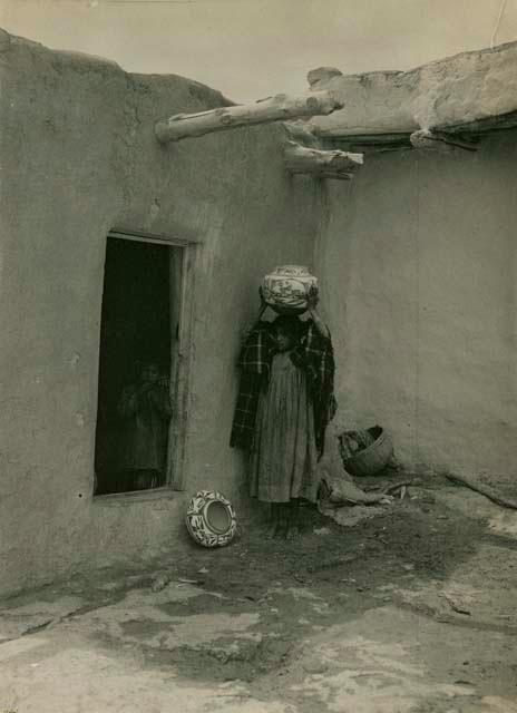 Tewa girl carrying ceramic jar on head, child in doorway