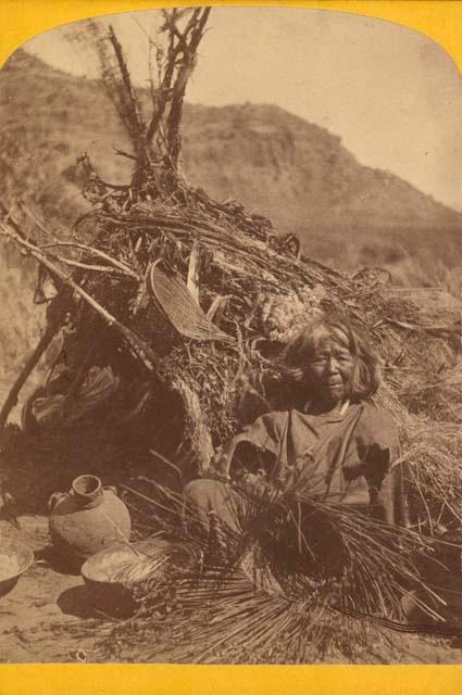 "The Basket Maker" - An old woman seated by a brush hut with baskets