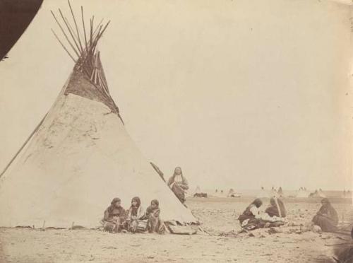 Arapaho camp; depicts three girls and man outside tent, others sitting in circle.