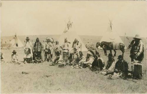 Group of men, seated, in a line; encampment is visible in background