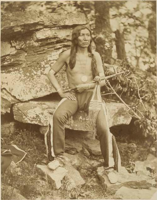 Man seated on rocks; he holds a pipe in his left arm