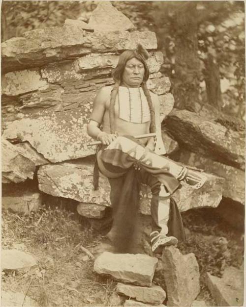 Man seated with legs crossed on rocks. He holds an axe in his right arm