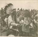 Man in headress and young child together amongst gathering of Crow Indians