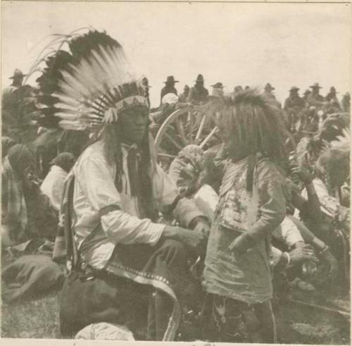 Man in headress and young child together amongst gathering of Crow Indians