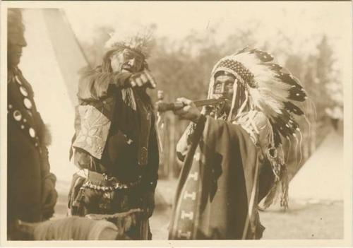 Man in headdress smoking pipe while other man points to its tip