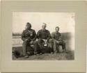 Three men, two White, one Native American, sitting on bench with field behind.