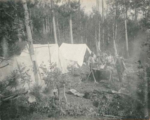 encampment with several A-frame tents and men outside eating and cooking