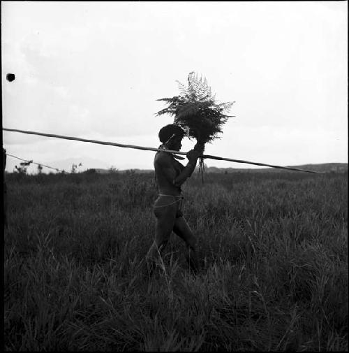 A warrior walks holding a long spear and a bunch of ferns