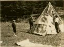 A boy watches as a man and woman lay bark strips over the frame of a Micmac wigwam