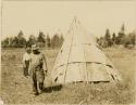 A man and woman, having put poles over the wigwam to hold down bark strips