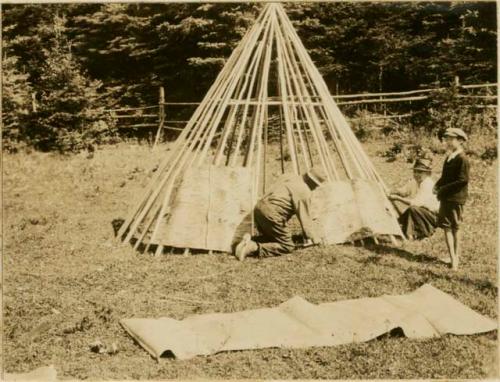 Man and woman laying on of covering bark strips over frame of Micmac wigwam