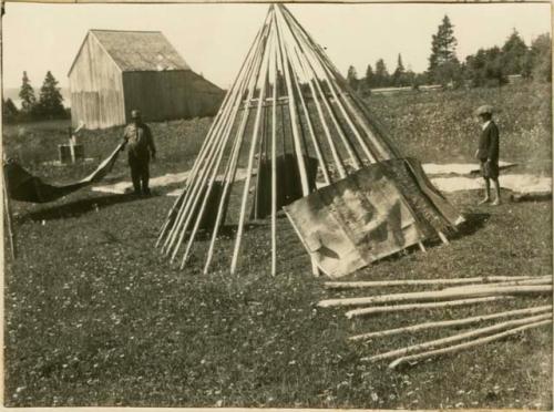 Boy watches as a man and woman lay bark covering over the frame of a wigwam