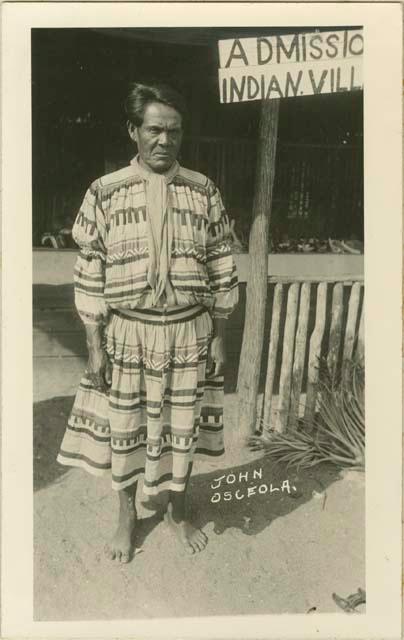 Photograph of John Osceola standing in front of "Admission Indian Village" sign