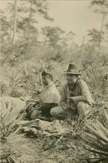 Seminole man and woman crouching amidst brush