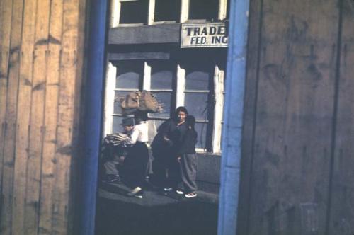 Looking through fence at people in front of building, one with bundle of sticks