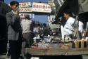 People at a street market, including stall keeper, potential customer, goods, and shop signs