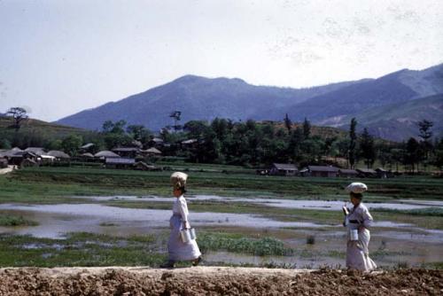 Two women walking by rice paddies, with sacks on their heads and carrying containers