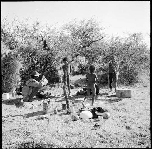 Man squatting in front of a skerm, with three children standing near him, with enamel pans and oil cans on the ground, boy playing with a toy car in the background