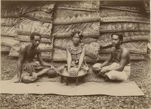 "Making Kava" - a woman and two men seated on mat, woman holds kava root and men hold bowls