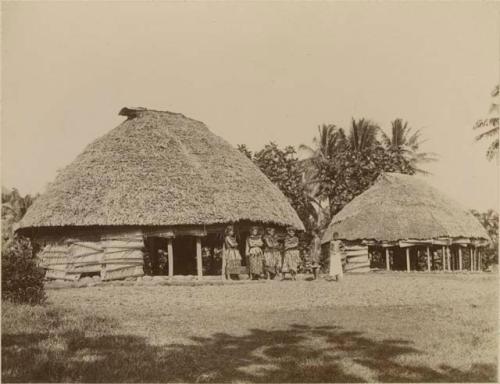 Four Samoan girls and two boys standing nearby two round houses or fale tele