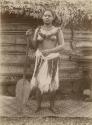 Samoan woman, posed, standing on mat holding a paddle in front of a house