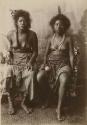 Two Samoan women sitting, one with scars on her leg holding a fan, the other holding a flower