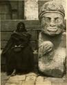 Native woman seated beside a large stone statue