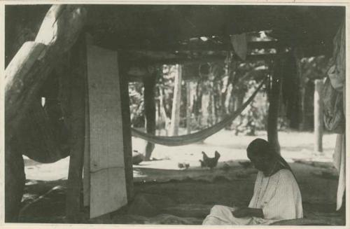 Woman sitting at loom inside structure, with chickens in background