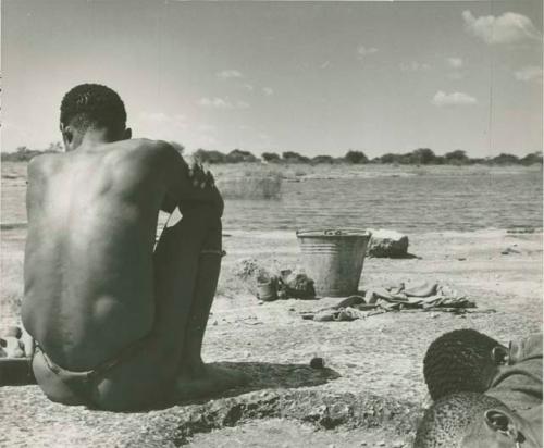 Man sitting at the edge of a pan, view from behind, with two men sleeping next to him, bucket on the ground in front of them