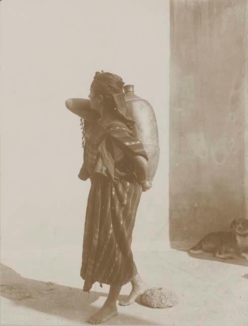 Woman carrying large vessel, with dog in background