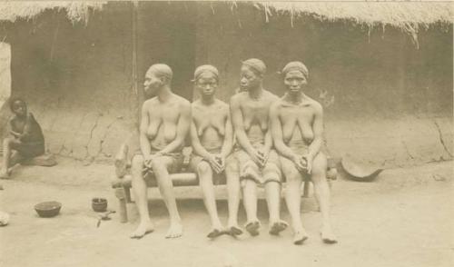 Four women with plaited hair sitting on a bench in front of a hut