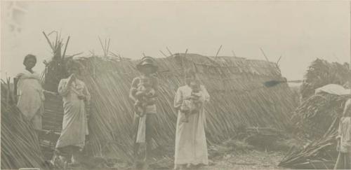 Three women, man and two babies in front of a hut