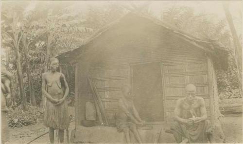 Two men and a woman in front of hut