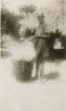 Man with large drum, basket or ceramic vessel in front of a hut