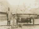 Man standing next to object tied to a post, with huts in background