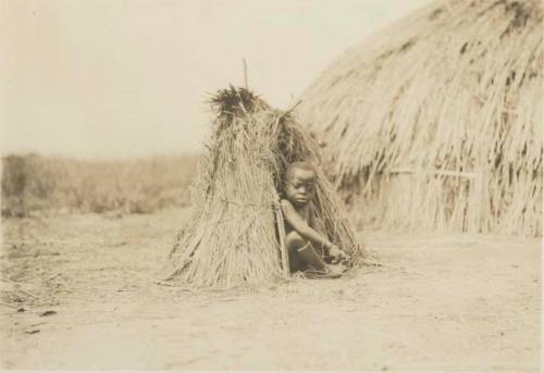 Child sitting under small hut, larger hut in background