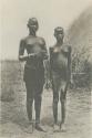 Two girls standing, with hut in background